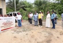 Hope Beyond Addiction: Caritas Freetown Empowers Kush-Affected Youth Through REAR Rehabilitation Project Group of people outdoors on a dirt yard holding a banner that welcomes Archbishop Edwards Tamba Charles, near a building and vehicle in the background, sunset-like daylight.