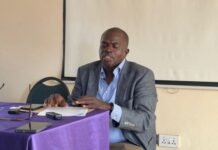 CSO Consortium Endorses Gento’s Banana Island Port, Predicts Massive Job Creation and Economic Growth Man in a gray blazer and blue shirt reads from papers at a table with a purple cloth, speaking softly into a discussion. It appears to be a small meeting or presentation.