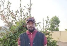 Dr. Tonya Musa Calls for New Social Contract as Sierra Leone’s Press Freedom Declines Man standing outdoors in a gravel lot, wearing a dark utility vest over a maroon polo, a cap, and a gold watch.
