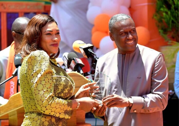 Woman in a gold sequin dress hands a glass award to a smiling man at an outdoor press event with microphones nearby.