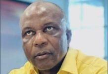 NASSIT DG Turns 60…It’s Time To Go Portrait of a man with a shaved head wearing a yellow shirt, looking slightly upward, in a bright indoor setting. The shirt has a blue logo on the chest.