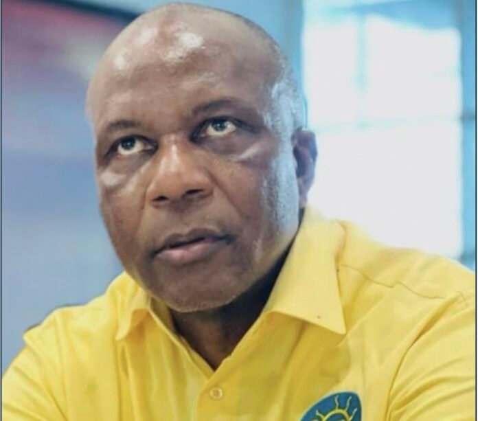 Portrait of a man with a shaved head wearing a yellow shirt, looking slightly upward, in a bright indoor setting. The shirt has a blue logo on the chest.