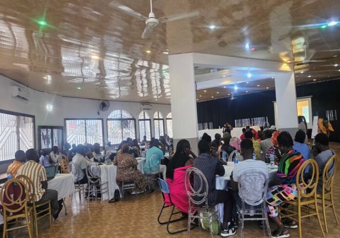 Large hall filled with people seated at round tables, listening to a speaker on a stage at the front.