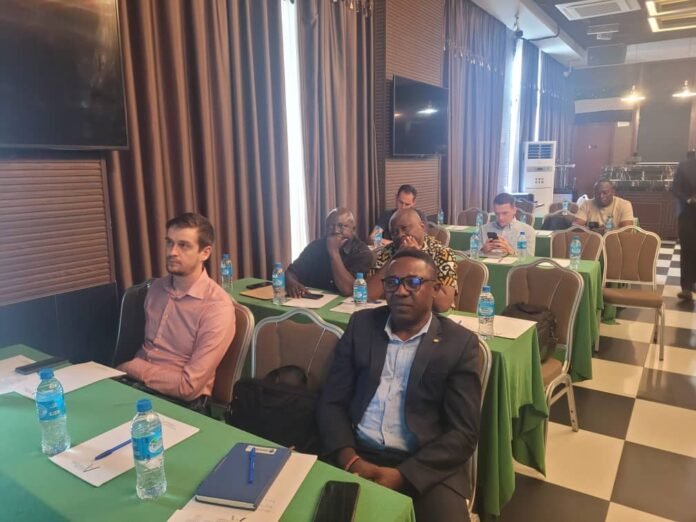 Participants at a conference seated at tables with green tablecloths, water bottles and notebooks on them in a banquet-style room.