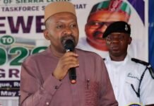VP Urges Discipline and Unity at Pre-Departure Orientation as 800 Pilgrims Prepare for Hajj Man in brown traditional outfit and beige cap speaks into a microphone at a public event, with a banner behind him and a uniformed officer in the background.