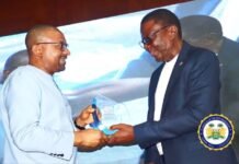 Vice President Bags Distinguished Award, Advances Workers’ Power for Sierra Leone’s Transformation on Labour Day Two men on stage exchange a glass award during a ceremony, one in a light gray suit and the other in a dark blazer, with a blue backdrop behind them.