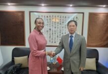 ICPNC Executive Secretary Engages Chinese Ambassador to Deepen Cooperation on National Cohesion Two professionals shake hands in a formal office; woman in a pink dress and a man in a gray suit, with calligraphy art behind them.