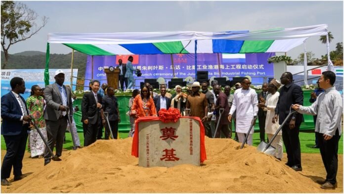 Officials and locals gather around a ceremonial stone on a sand mound at a port groundbreaking under a white-and-blue canopy.