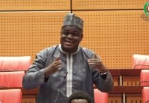 Sierra Leonean Lawmaker Raises Concerns Over President Barrow’s Third-Term Bid at ECOWAS Parliament Man in traditional gray attire and patterned hat speaks at a microphone in a legislative chamber, gesturing with his hands.