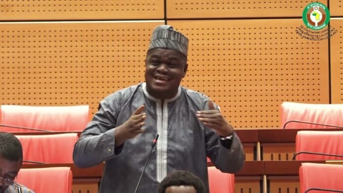 Man in traditional gray attire and patterned hat speaks at a microphone in a legislative chamber, gesturing with his hands.