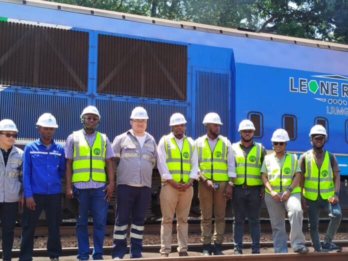 Group of railway workers in safety vests and hard hats standing on tracks in front of a blue locomotive.