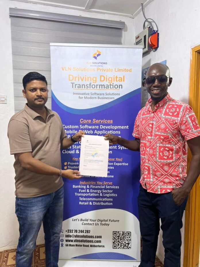 Two men stand indoors, one presenting a certificate to the other in front of a VLN Solutions banner that reads 'Driving Digital Transformation'.