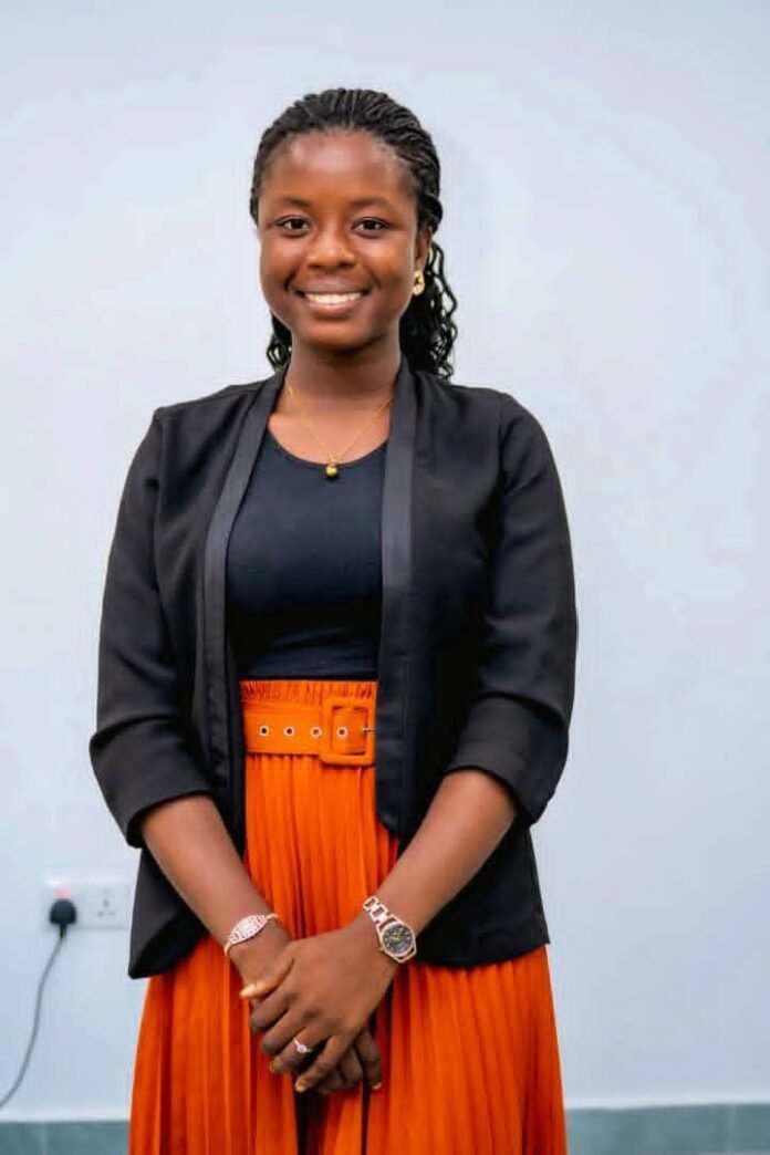 Young woman standing with folded hands, smiling, wearing a black blazer, black top, and bright orange skirt in a neutral setting.