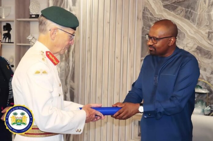 Senior officer in a white uniform presents a blue rolled document to a man in dark blue attire during a formal ceremony, with a VP emblem nearby.