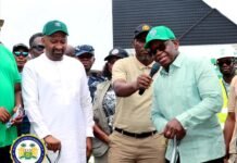 VP Juldeh Jalloh Joins President Bio for Landmark SLPP Regional Office Groundbreaking in Kenema Two men stand at a public event: one in white traditional clothing and green cap, the other in a light green shirt with a microphone, as onlookers watch and a government seal appears nearby.