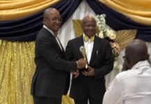 Kobi Walker Honoured for Outstanding Corporate Leadership at 2026 West Africa Heroes Awards Two men in suits on a stage exchanging a trophy during an award ceremony, photographer capturing the moment.