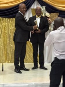 Two men in suits on a stage exchanging a trophy during an award ceremony, photographer capturing the moment.