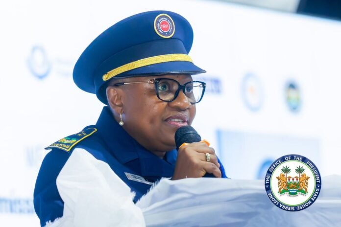 Female public official in a blue uniform and cap speaks into a microphone at a podium with a government seal visible near her.