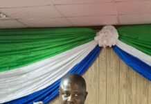 Attorney General Says Proposed Constitution Will Shift Sierra Leone From “Survival to Prosperity” Man in a dark suit giving a speech at a lectern decorated with green and white fabric, holding a microphone and gesturing with his left hand.