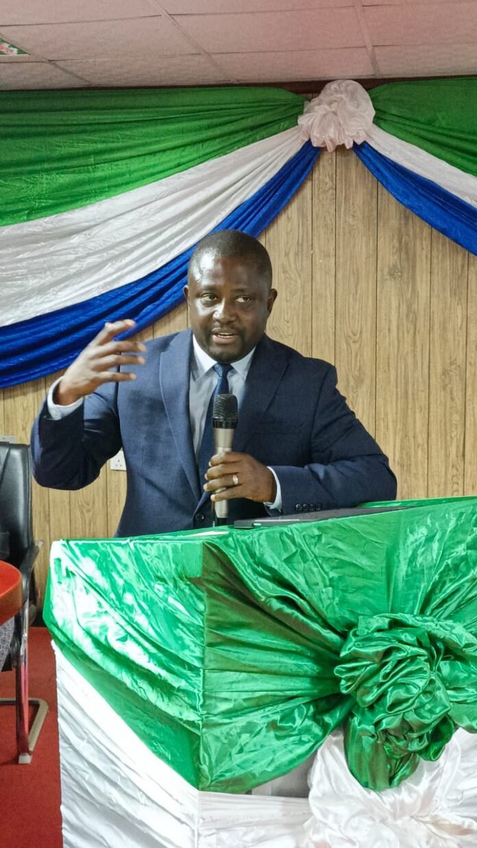 Man in a dark suit giving a speech at a lectern decorated with green and white fabric, holding a microphone and gesturing with his left hand.