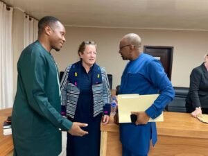 Three professionals in a meeting around a wooden table: a man in green scrubs, a woman in a striped jacket, and a bald man in blue holding folders and a laptop.