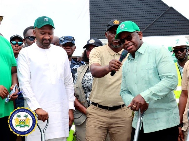 Group of men at an outdoor event; a man in a green cap speaks into a microphone while another in white traditional clothing stands beside him; government seal visible in the lower left corner.