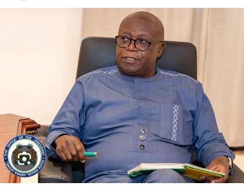 Man wearing a blue traditional outfit sits in a chair, holding a green pen and a notebook in a formal setting; official seal visible bottom-left.