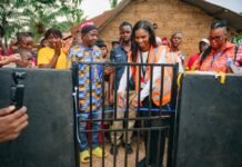 SLCAA DG Donates Personal Water Well to Royeama Village, Restores Hope After 30 Years of Water Crisis Woman in an orange safety vest opens a metal gate as a diverse crowd watches in a rural outdoor setting.