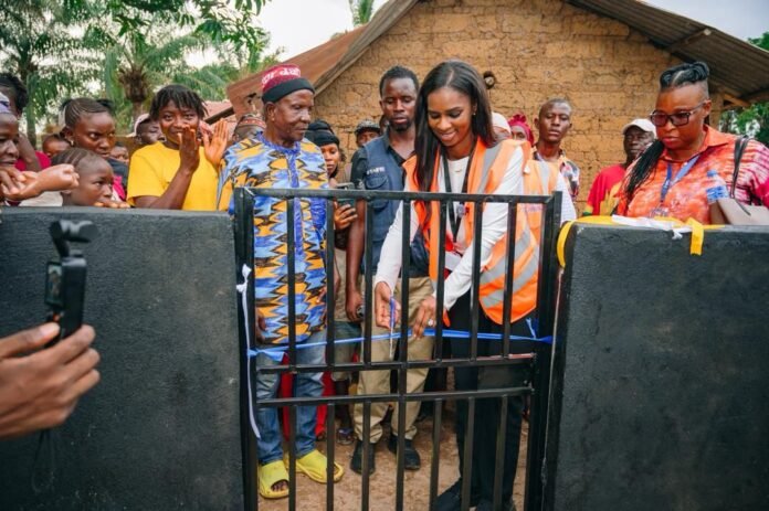 Woman in an orange safety vest opens a metal gate as a diverse crowd watches in a rural outdoor setting.