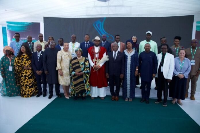 Group of diverse dignitaries and attendees posing on stage at a formal event, with a central figure in red traditional attire flanked by officials in suits and dresses.