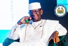 President Bio Hosts Leadership Colloquium, Calls for Bold Youth Action Across Africa Man in white traditional attire and cap, speaking into a green-tipped microphone on stage; a seal is seen in the backdrop.