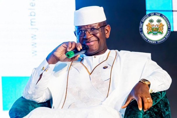 President Dr. Julius Maada Bio Man in white traditional attire and cap, speaking into a green-tipped microphone on stage; a seal is seen in the backdrop.