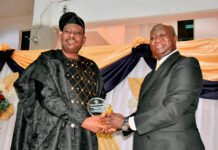 Ambassador Justice Desmond Babatunde Edwards Receives Prestigious 2026 West Africa Heroes Award Two men in formal attire shake hands as one receives a glass trophy at an awards ceremony in front of orange and navy drapes.
