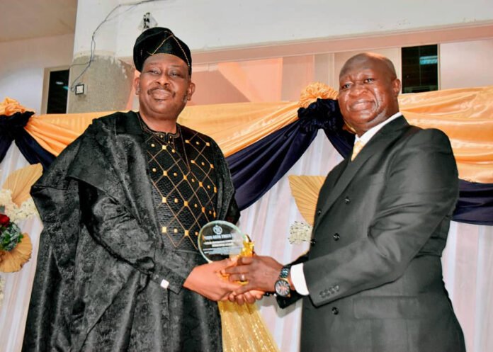 Two men in formal attire shake hands as one receives a glass trophy at an awards ceremony in front of orange and navy drapes.