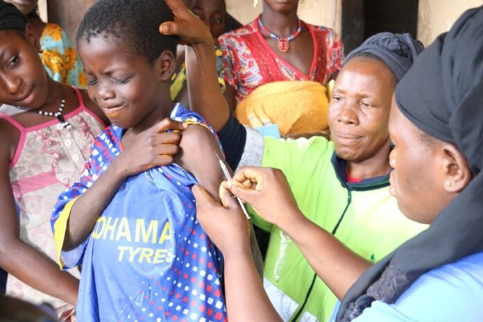 Young person receiving a vaccine shot in the upper arm from a healthcare worker, with a group of community members watching nearby.