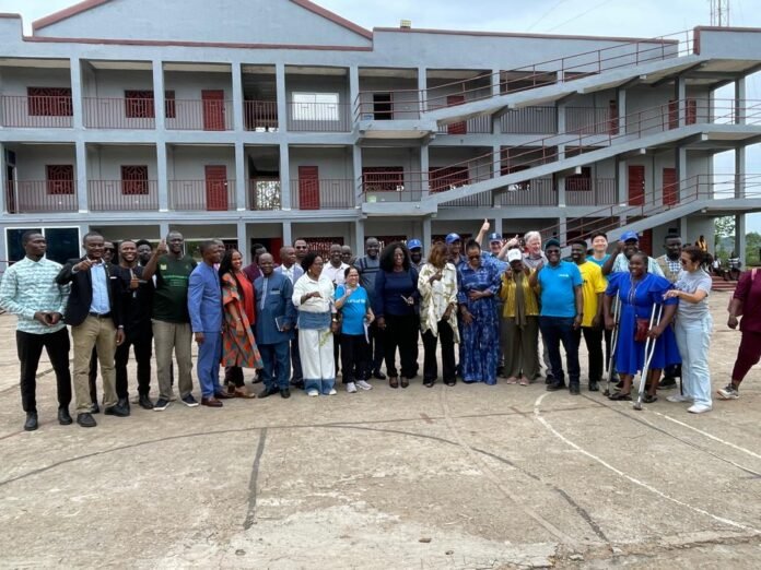 Group of diverse people standing together for a photo in front of a gray building with red doors and external staircases, outdoors on a paved area, suggesting a community event or gathering.