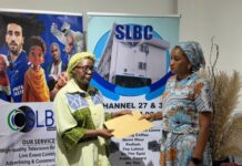 Orange Sierra Leone Supports SLBC with Financial Donation Following Fire Incident Two women stand in front of SLBC banners as one hands a yellow envelope to the other during a formal exchange at a media event.