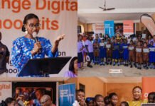 Orange Foundation Rolls Out First Digital Learning Kits to Regent Square School in Freetown Woman in a blue floral dress speaks at a podium during a school event, with a large group of students holding tablets in the background.