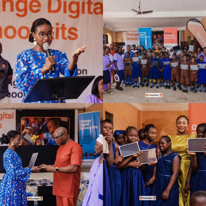 Woman in a blue floral dress speaks at a podium during a school event, with a large group of students holding tablets in the background.