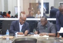 Government, Contractors Commit Over US$4.2 Million to Finalize Stalled Hospital Projects Two men in business attire sign documents at a conference table as colleagues observe in a meeting room; water bottle and notebooks nearby.
