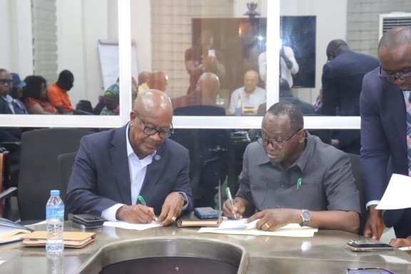 Two men in business attire sign documents at a conference table as colleagues observe in a meeting room; water bottle and notebooks nearby.