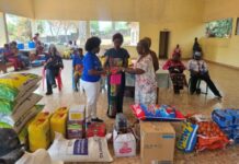 Rotary Club of Freetown Supports King George VI Memorial Home for the Aged with Essential Supplies Community aid distribution in a large hall: volunteers hand a package to two women while others sit or wait among stocked supplies (bags of rice, oil, and boxes) for a relief event.