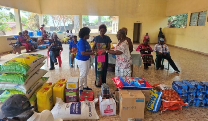 Community aid distribution in a large hall: volunteers hand a package to two women while others sit or wait among stocked supplies (bags of rice, oil, and boxes) for a relief event.