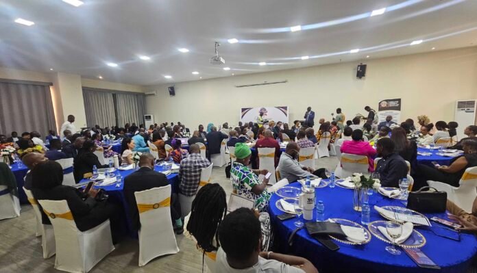Large banquet hall with round tables covered in blue cloths and many formally dressed attendees listening to a speaker at a podium on a stage.",