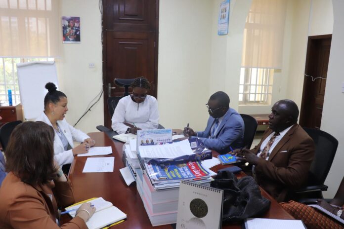 Five professionals in a meeting around a cluttered conference table with documents and magazines on display in a bright office setting.