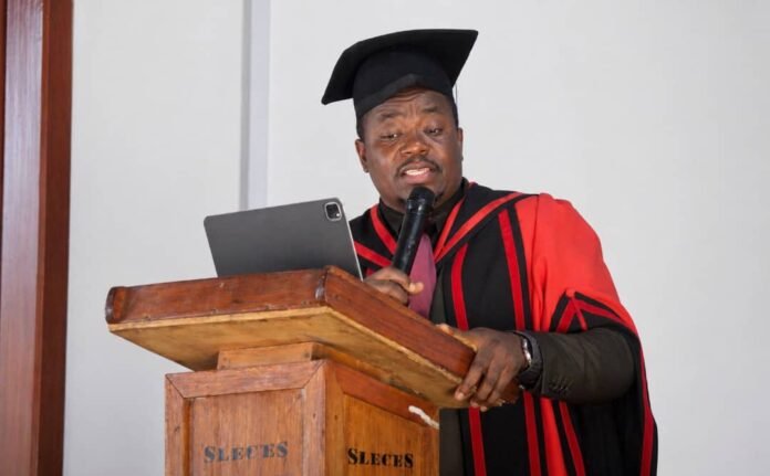 Graduating speaker in red and black gown delivers a speech at a wooden lectern, holding a microphone and tablet.