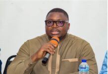 First Batch of 300 U.S. Deportees Arrives in Freetown Man wearing brown traditional attire speaks into an orange microphone at a panel, with a water bottle on the table nearby and a pale background.