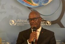Dr. Austin Demby Highlights Sierra Leone’s Health Reforms at Global Health Summit Man in a dark suit and glasses speaks into a handheld microphone at a conference backdrop with Africa CDC logos.
