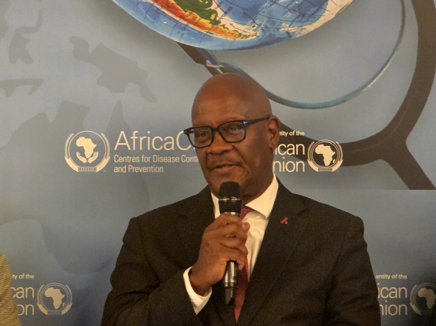 Minister of Health, Dr Austin Demby Man in a dark suit and glasses speaks into a handheld microphone at a conference backdrop with Africa CDC logos.