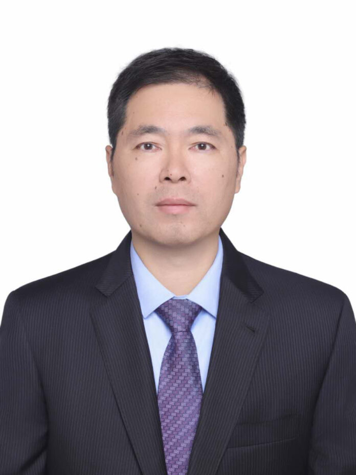 Professional headshot of a man in a dark suit, light blue shirt, and purple tie, looking at the camera against a white background.